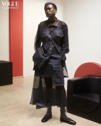 Model wearing dark denim jacket and sheer skirt ensemble holding a matching handbag in a modern interior setting