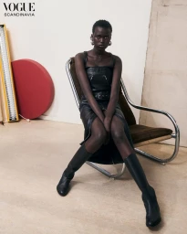Model seated in a chrome armchair wearing a black leather dress and knee-high boots in a minimalist studio.