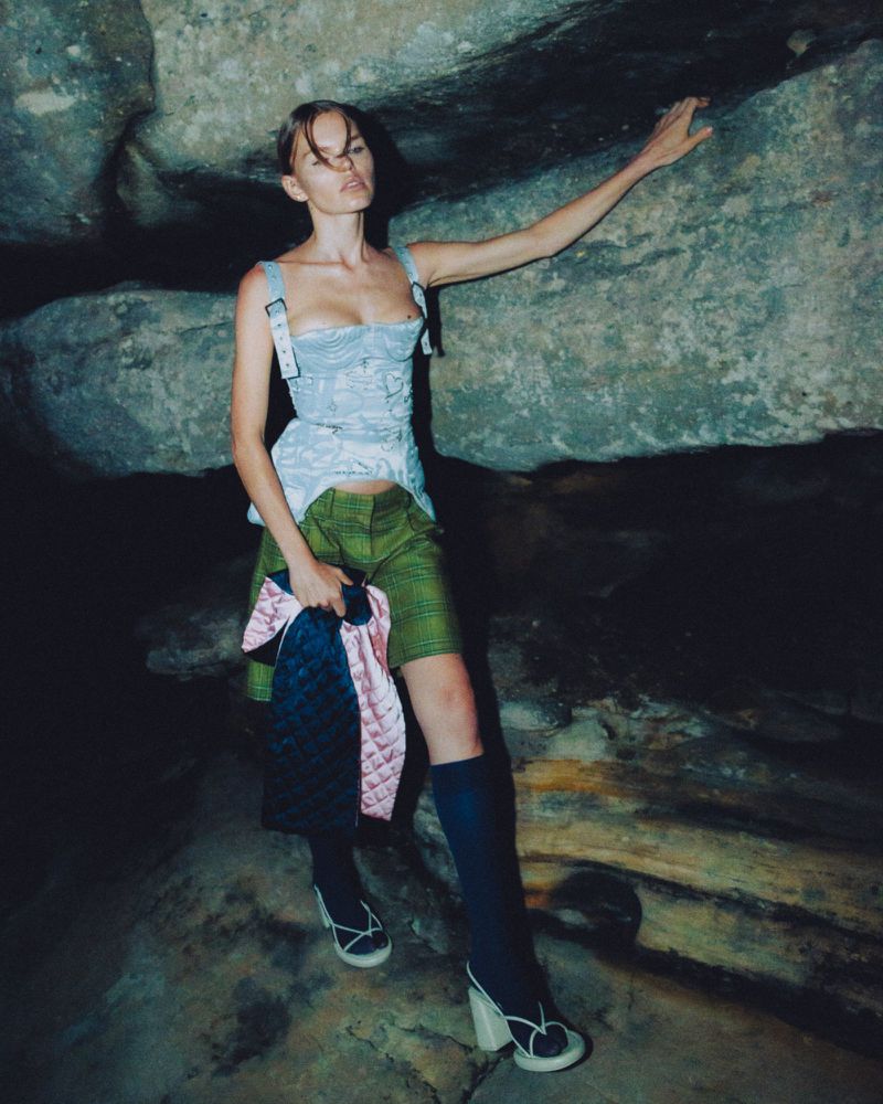 Adult female model poses in corset, shorts, socks, heels with stone cave background, holding patterned handbag. Editorial fashion photography.