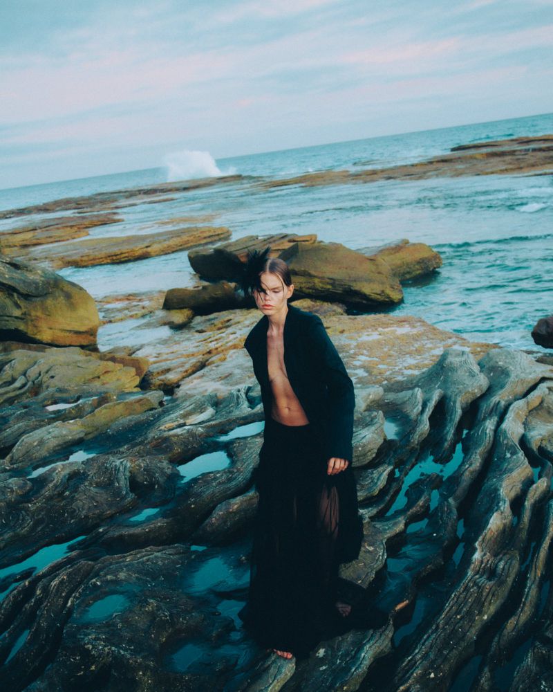An adult stands on a rocky beach wearing a dark coat and sheer skirt, the ocean and sky visible in the background on an overcast day.