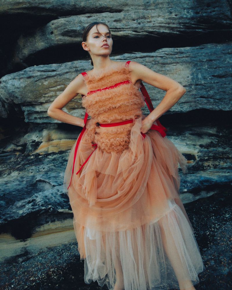 A fashionable adult female model poses in a layered peach tulle dress with red accents against a textured stone background in a portrait-style shot.
