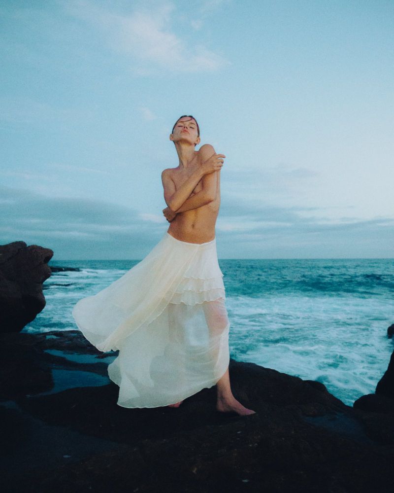 Woman in flowing white skirt stands on rocks near the ocean, arms crossed, basking in light. Sea and sky backdrop create serene scene.