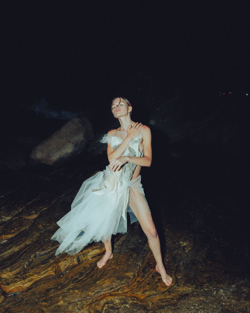 Woman poses in a light-colored dress on rocky beach at night. The image features a dark background and highlights the dress's flow and texture.