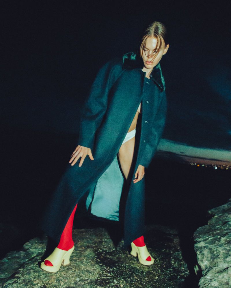 Fashion model in a teal coat and red socks standing on a rock at night with city lights in the distance, posing for a studio portrait.