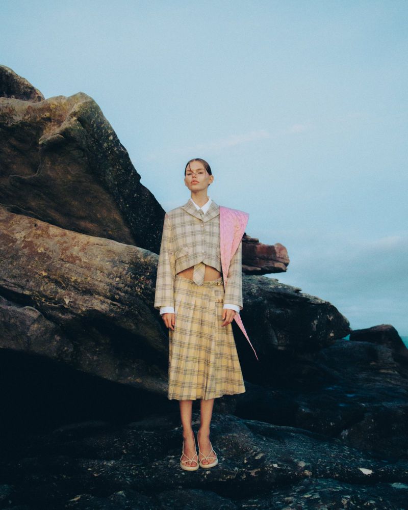 A fashionable woman in a plaid blazer and skirt poses on a rocky landscape against a bright sky with a large rock formation to the left.