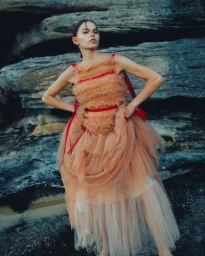 Model wearing a layered peach tulle dress with red ribbon straps posing outdoors against a rocky stone cliff background.
