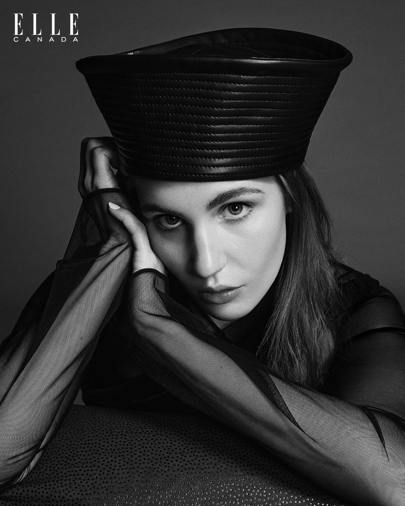 Black and white portrait of a woman with straight hair wearing a black hat and top, hands at face, gazing into the camera.