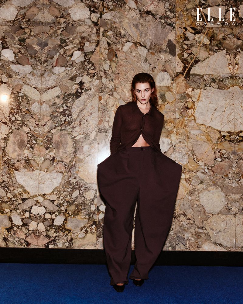 Fashion editorial shot of a brunette adult female model in a sheer blouse and oversized pants, posing in front of a marble wall.