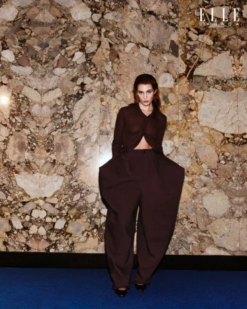 Fashion editorial shot of a brunette adult female model in a sheer blouse and oversized pants, posing in front of a marble wall.