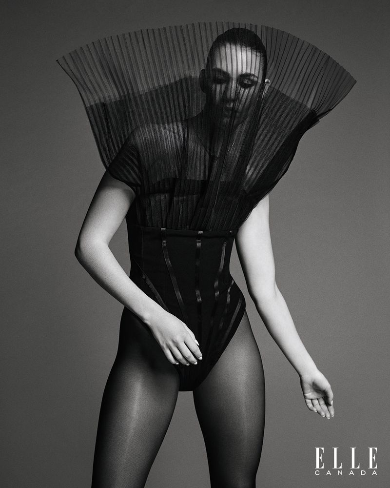 A woman in a black bodysuit with sheer pleated fabric detail stands against a gray backdrop in a studio fashion shot for ELLE Canada.