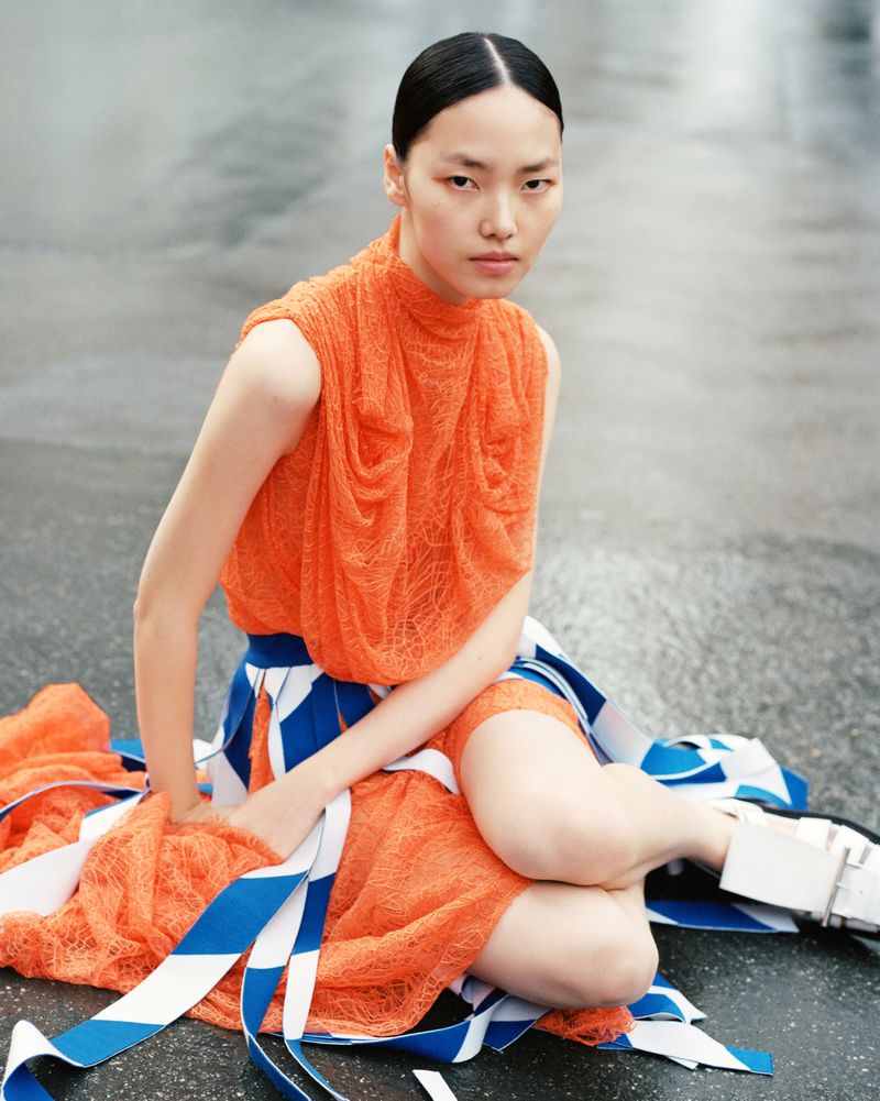 Asian fashion model poses in orange top and blue/white skirt on grey pavement, portrait orientation, direct gaze, beauty and fashion shoot.