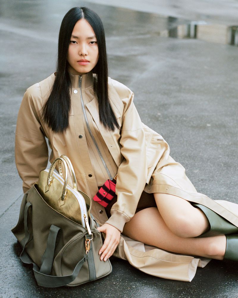 Young Asian woman poses seated with olive-green duffel and metallic handbag, wearing a beige jacket and green boots against wet grey concrete.