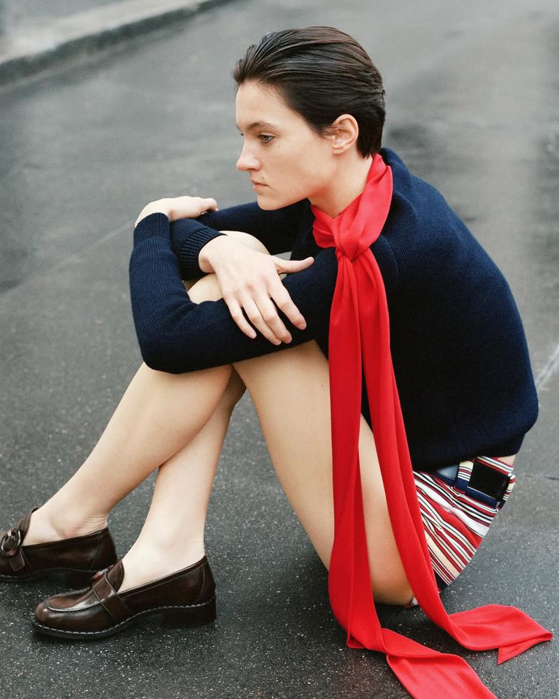 Woman sitting on the ground wearing a navy sweater, striped shorts, brown loafers, and a long red scarf with her arms resting on her knees.