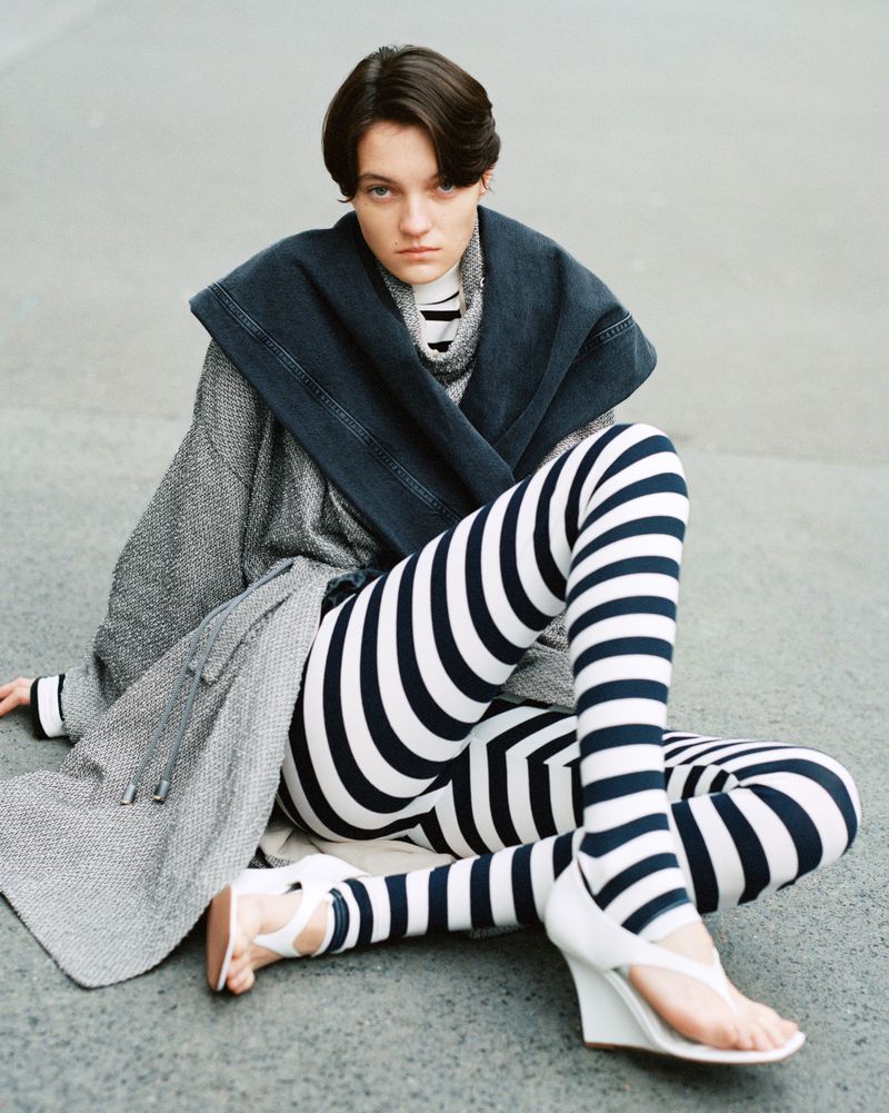Woman in striped leggings and white sandals sits on concrete. She wears a gray coat and dark blue scarf in a casual setting.