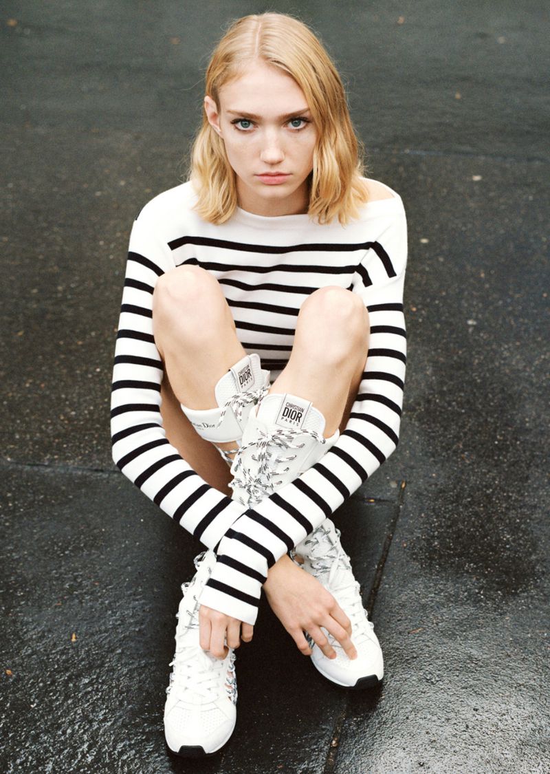 A blonde adult woman sits outside on a wet surface, wearing a striped sweater and white sneakers. The shot is from a high angle.