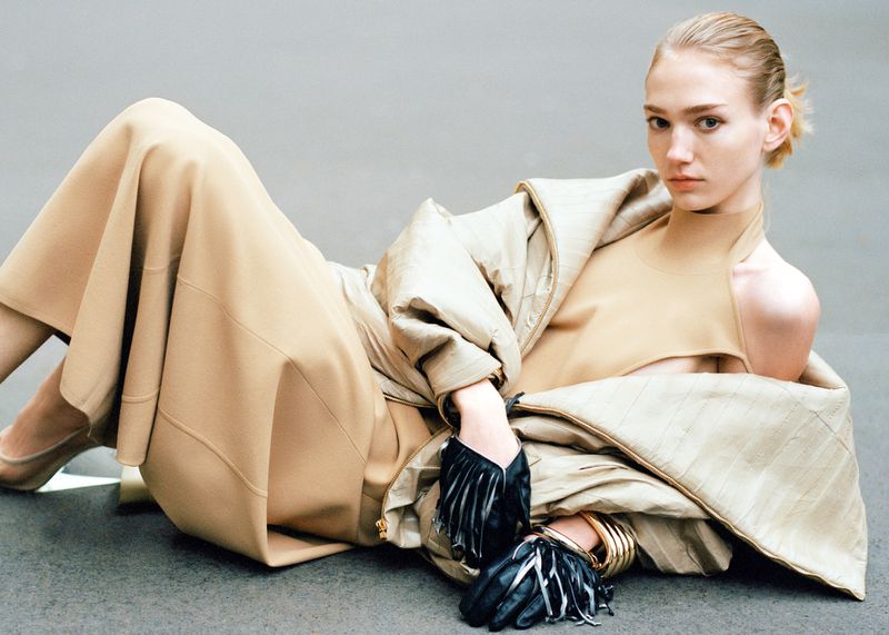 Fashion portrait of a blonde female model lying on a neutral background wearing a tan outfit, jacket, and black fringe gloves.