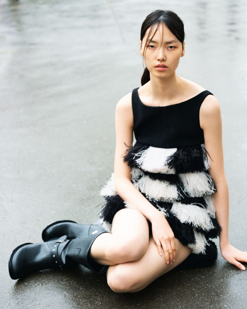 Young woman in a sleeveless, feathered dress poses on a wet surface wearing black boots, creating a striking fashion editorial image.