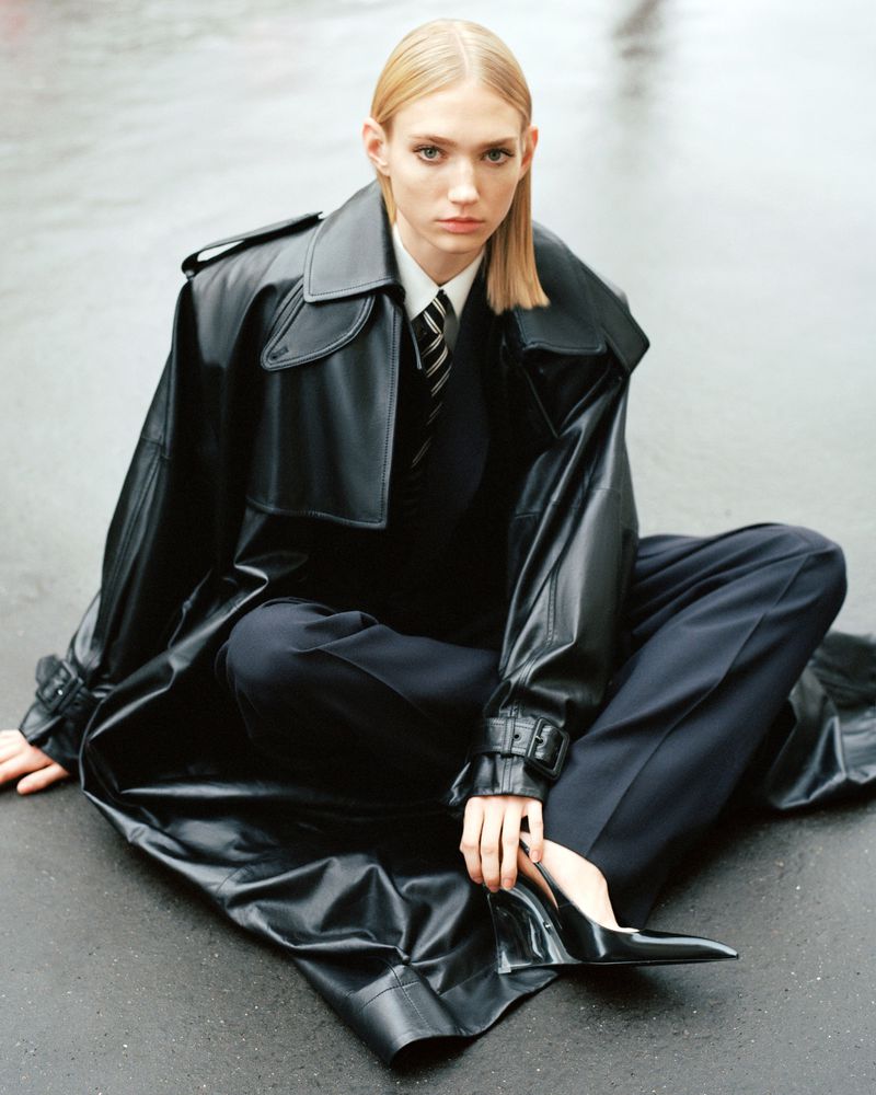 A slender blonde woman in a black trench coat, white shirt, and tie sits cross-legged on pavement, gazing directly at the camera.