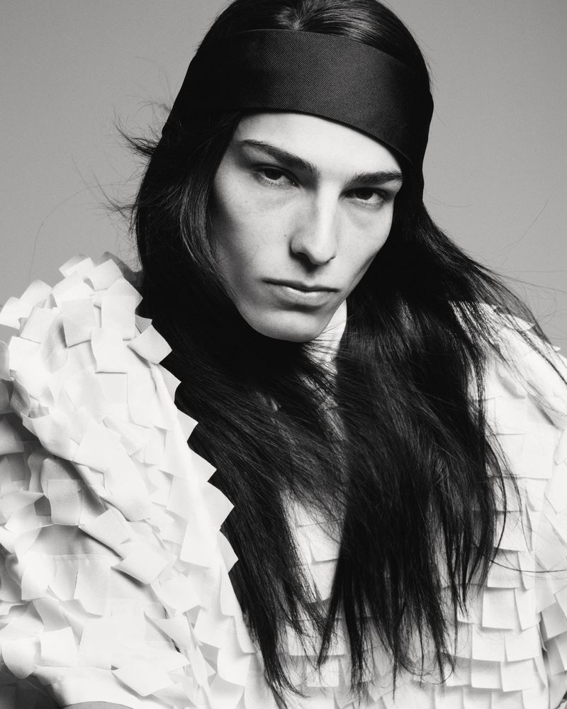 Monochromatic portrait of a model with long dark hair and a black headband, wearing a textured white top against a neutral backdrop.