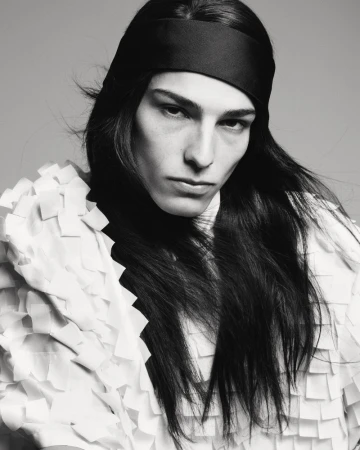 Monochromatic portrait of a model with long dark hair and a black headband, wearing a textured white top against a neutral backdrop.