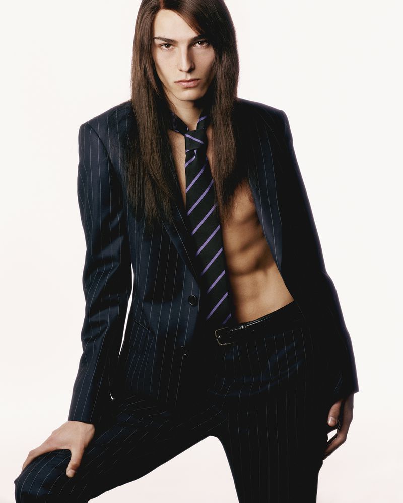 A model poses indoors wearing a pinstripe suit jacket and matching pants, with a striped tie, showcasing a fashion-forward style.