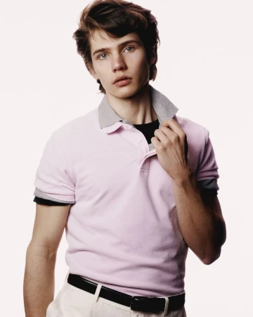 Portrait of a young adult male in a pink polo shirt with a grey collar, wearing white pants and a black belt against a white background.