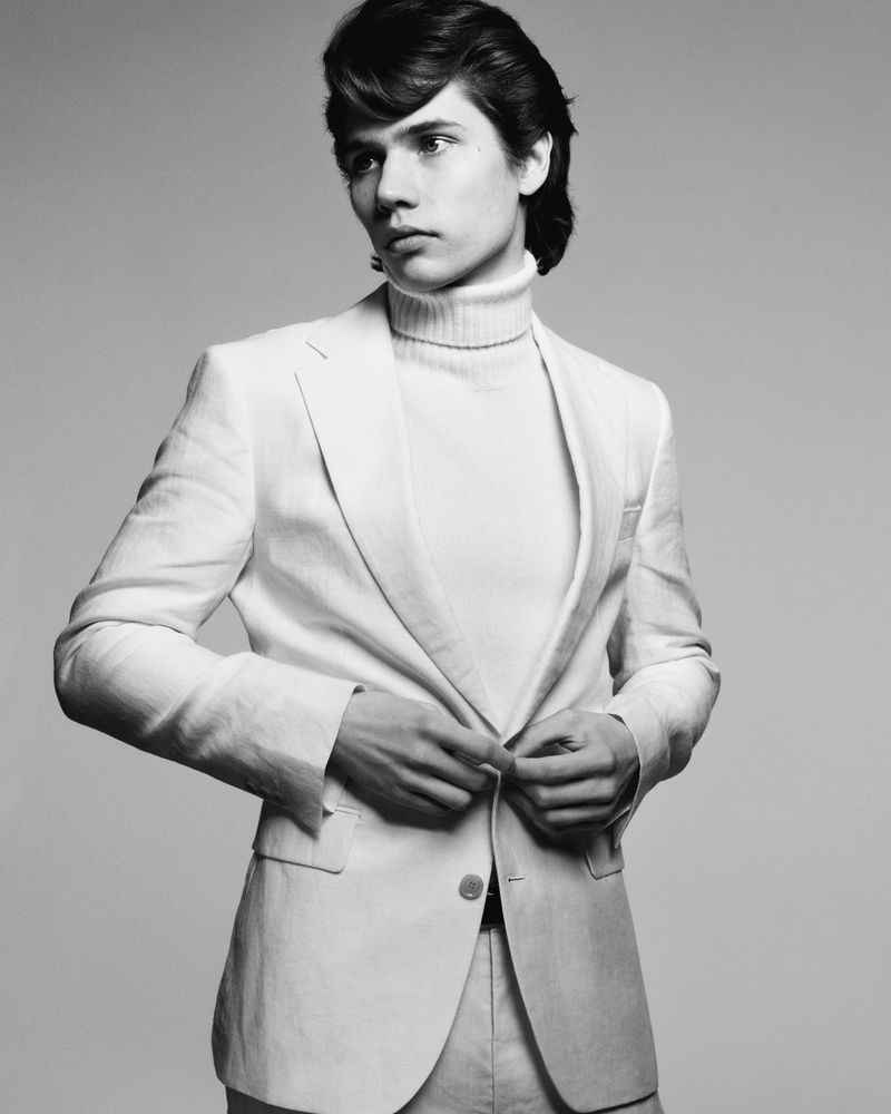 Monochrome portrait of a young adult male in a white linen suit and turtleneck sweater, looking off-camera in a photo studio setting.