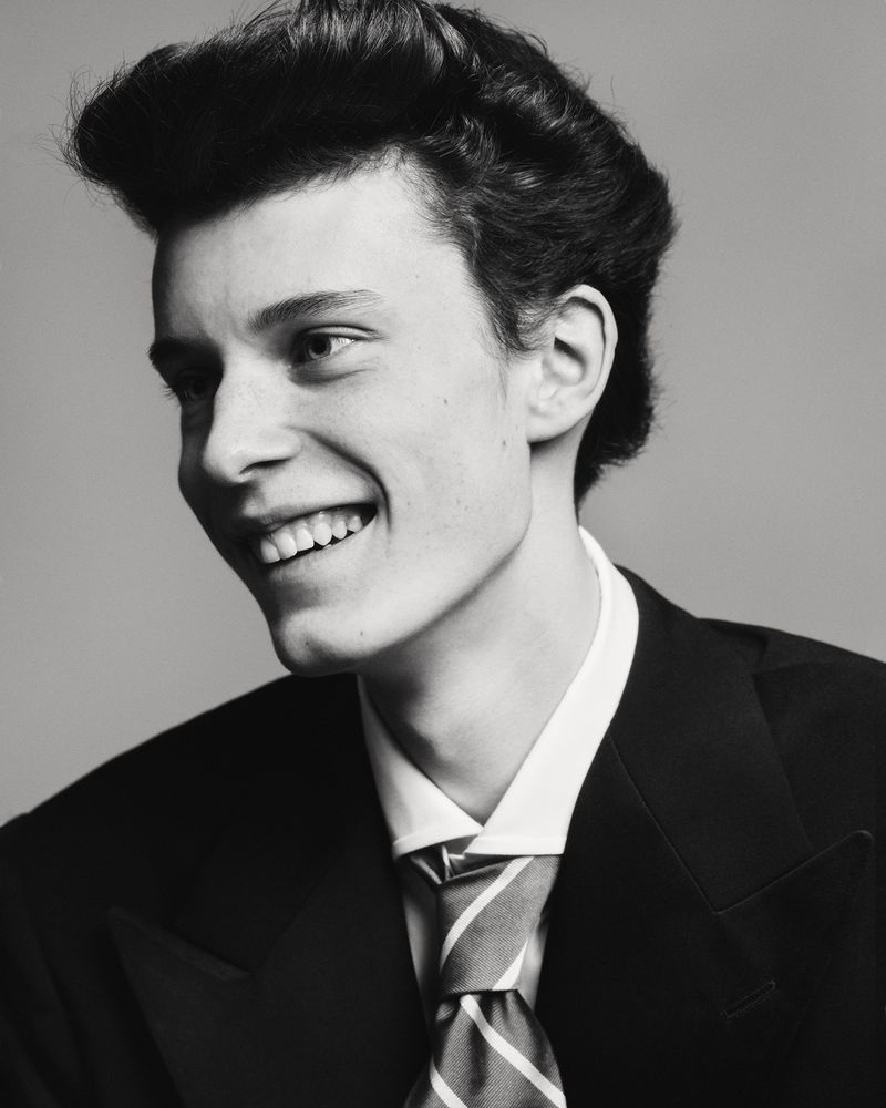 Black-and-white portrait of a smiling young man in a suit and tie, his hair styled in a retro fashion, set against a plain backdrop.