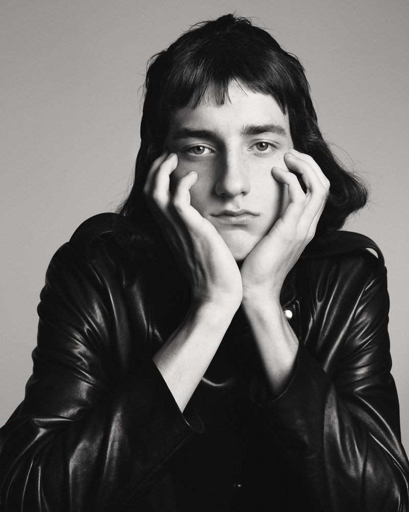 Close-up portrait of a young adult with shoulder-length hair, wearing a black leather jacket, posing with hands framing their face against a grey background.