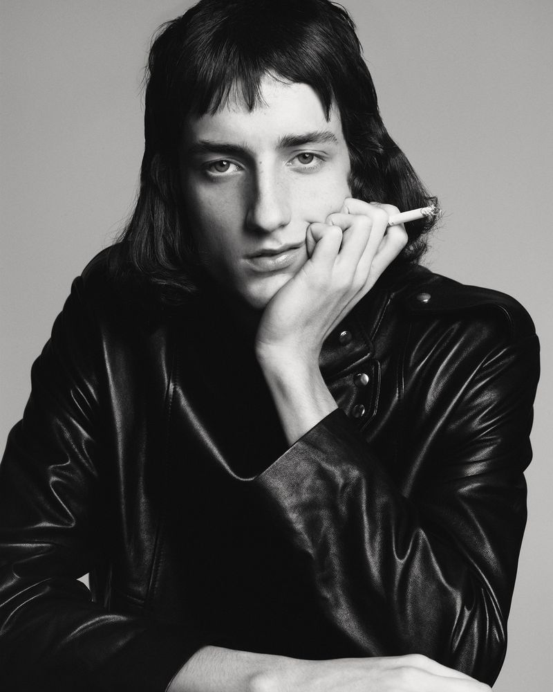 Monochrome portrait of a young adult with long dark hair, smoking, wearing a black leather jacket against a neutral studio backdrop.