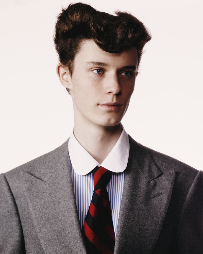 Portrait of a young fair-skinned man in a grey suit jacket, striped shirt, and red-striped black tie against a white backdrop.