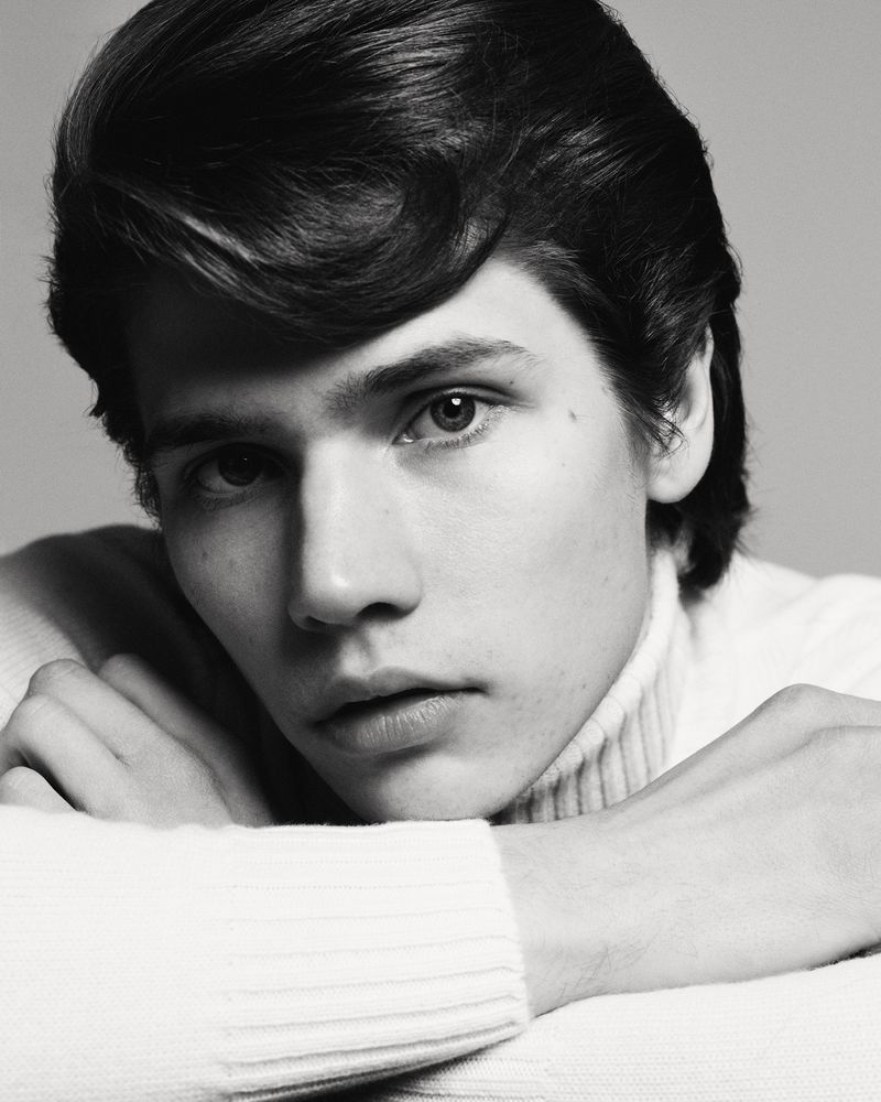 Black and white portrait of a young man with dark hair in a white turtleneck sweater, posing indoors with his arms resting on a surface.