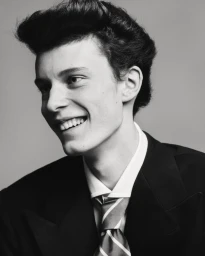 Portrait of a smiling young male model wearing a dark suit jacket, white shirt, and striped tie against a plain grey studio backdrop.
