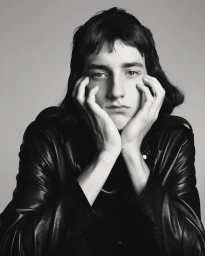 Portrait of a young male model with dark hair wearing a black leather jacket against a grey background.