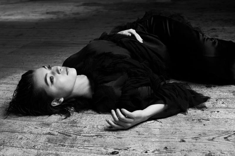 Monochrome portrait of a woman lying on her back on a distressed wood floor, wearing a dark, textured dress and looking upwards.