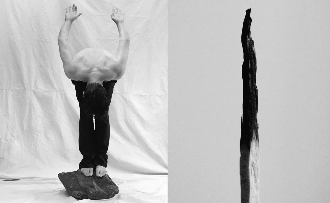 Monochrome photo of a man balancing on a rock, arms raised overhead, juxtaposed with a vertical piece of burnt wood against a bright white backdrop.