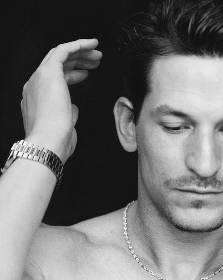 Close-up monochrome portrait of a man touching his hair, wearing a metal link watch and silver chain necklace.