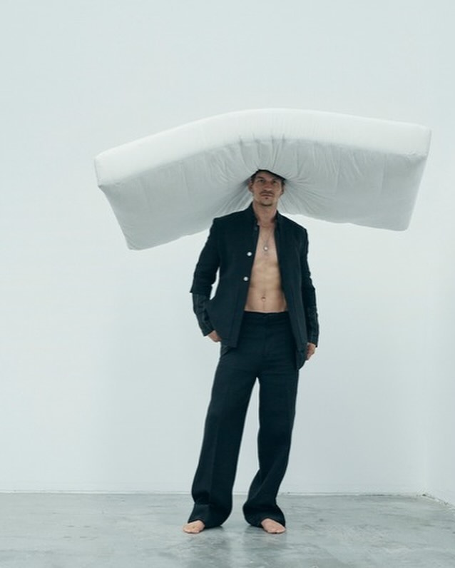 A man stands in a photo studio wearing a black suit, pants, and pillow on his head. He is standing barefoot against a white background.