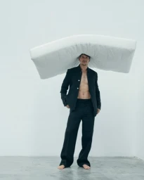 Man wearing a black jacket and trousers balancing a large white mattress on his head against a white background