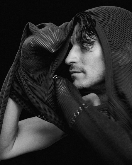 Black and white portrait of a man with dark hair, partially covered by a hooded mesh fabric, against a stark black background.
