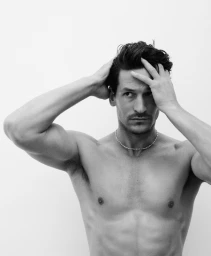 Black and white portrait of a shirtless man with dark hair running his hands through his hair against a plain white background.