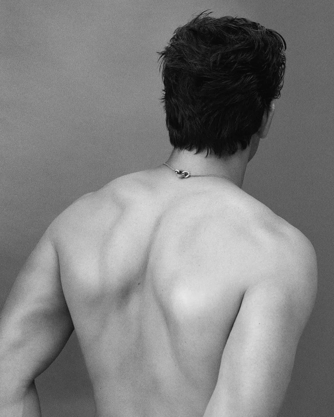 Back view of muscular adult male with short dark hair wearing a chain necklace, set against a plain grey studio backdrop.