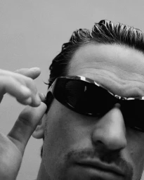 Close-up portrait of a man wearing dark sunglasses and adjusting them by his ear against a neutral background.