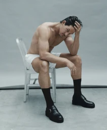 Shirtless man sitting on a white chair wearing white shorts, black socks, and black shoes against a grey studio backdrop.