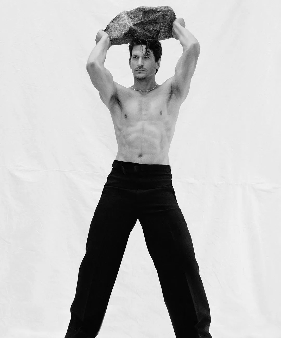 A shirtless adult male is lifting a stone overhead in a studio with a white background. He is wearing black pants and has dark hair.