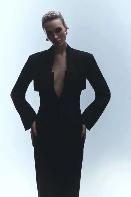 Fashion shot of an adult female model wearing a black dress with long sleeves, hands on hips, and stud earrings, against a white backdrop.