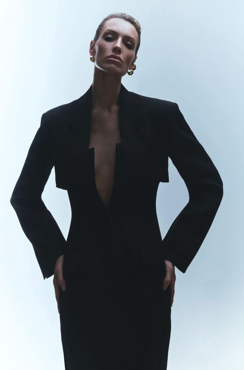 Woman in a black fitted dress and structured jacket stands in a brightly lit studio, presenting a high-fashion look with confidence.