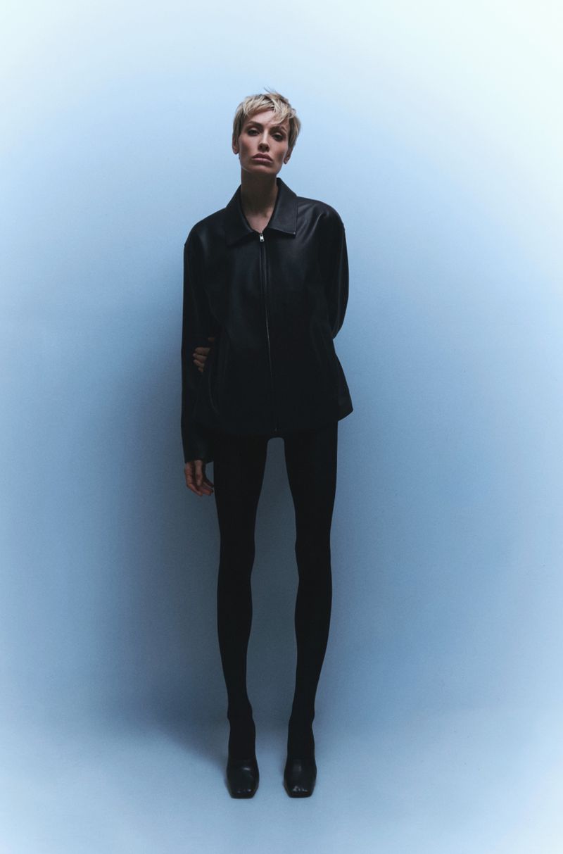 A blonde woman in a black jacket, leggings, and high heels stands in a photo studio with a light blue background. She is looking at the camera.