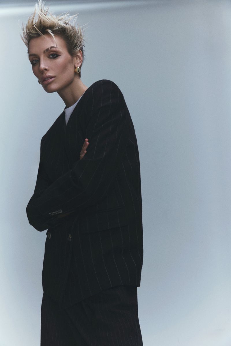 Adult woman in a black pinstripe suit jacket poses in a studio setting, looking towards the camera with a neutral facial expression.