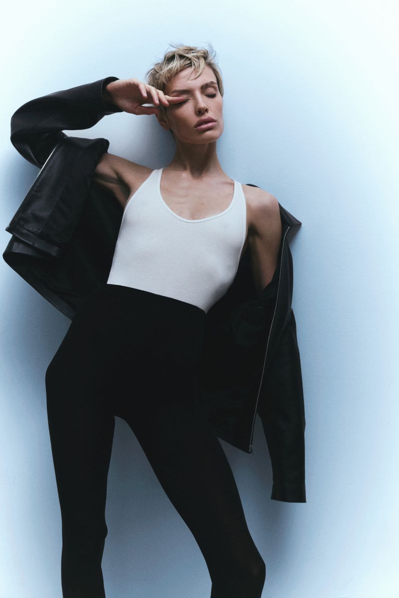 Wallis Day posing in a white tank top and black trousers with a dark jacket draped off her shoulders against a pale blue background.
